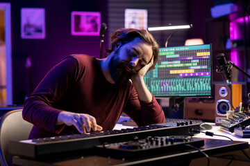 Tired artist losing sleep to learn new piano notes in home studio, playing midi controller electronic keyboard to compose songs. Producer being exhausted and stresses while he works on music.