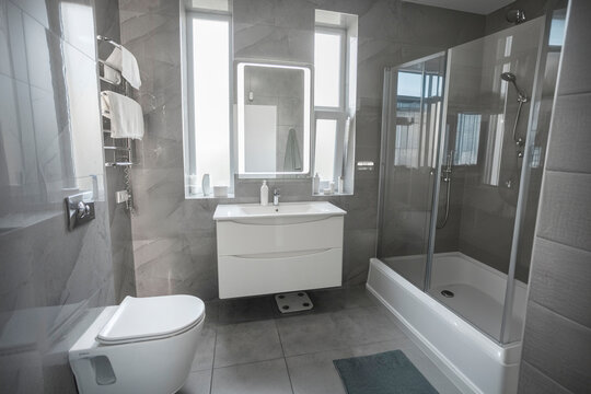 Minimalist bathroom interior design with grey tiles, mirror with led lighting and white furniture, washbasin, toilet, bathtub