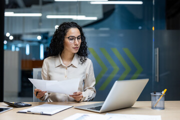 Confident businesswoman analyzing documents while using laptop in modern office. Casual attire suggests comfortable work setting. Emphasis on focus and productivity.