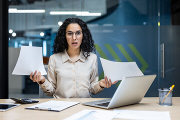 Confused businesswoman sits at desk holding documents in hands, surrounded by office tools like laptop and smartphone. She expresses uncertainty in modern office environment