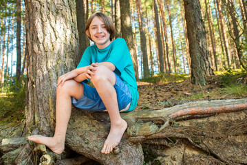 11 year old boy in shorts and a t-shirt, barefoot, sits on tree roots in a pine forest