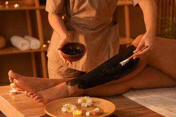 Massage therapist applying mud on woman's leg in dark spa salon, closeup