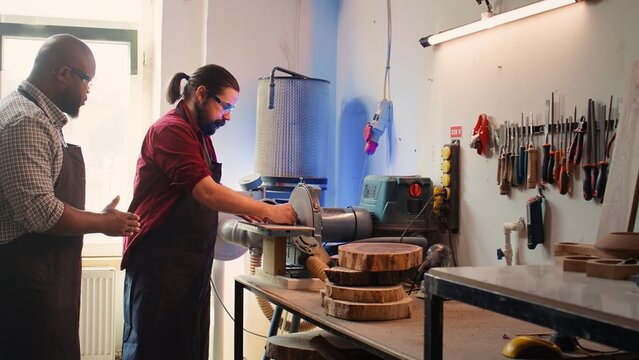 Man teaching african american trainee how to shape wooden bowl on disc sander in assembling shop. Worker learning from woodworking professional how to use grinding machine rotating disc, camera B