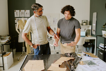 Curly haired designer holding fabric parts of future dress while mature tailor pointing out it