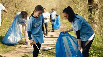 Mom and child collecting rubbish from the forest area using a long claw tool and garbage disposal bags. Little girl picking up trash and plastic waste with her mother, ecological justice. Camera A.