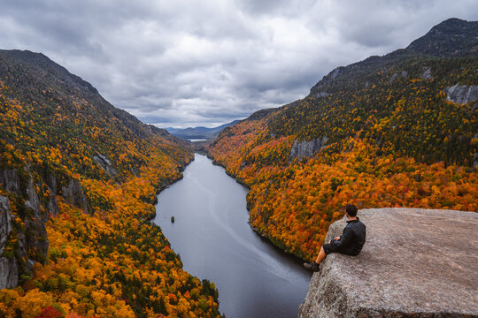 Hiker Sits on Edge of Mountain Indian Head Adirondacks New York in Autumn Peak Fall Foliage. Freedom Man Casually Looks Out Over Lake and Clouds