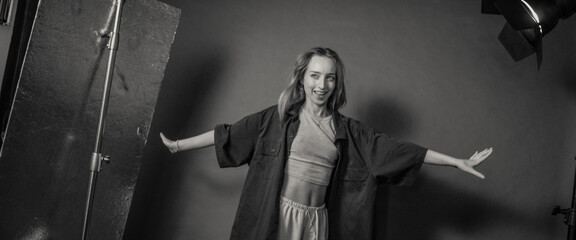 A woman stands in a studio, smiling with arms outstretched, dressed in casual attire, evoking a...