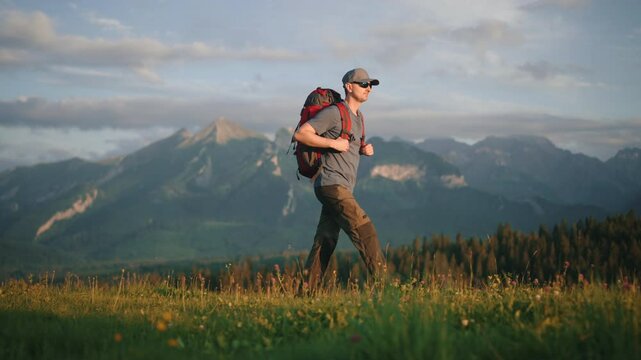 Side view following of man with red backpack and trekking poles walking on green grass meadow hill overlooking epic mountains covered with clouds. Male backpacker explorer hiking, sunny summer morning