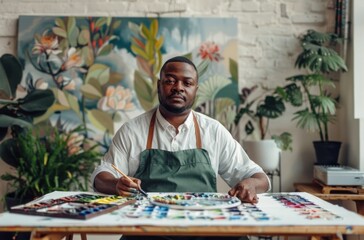 A Black male painter in a green apron focuses on his work, surrounded by lush floral paintings and a well-lit, plant-filled studio.