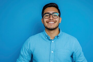 Fototapeta premium Portrait of young smiling latin man in blue shirt and glasses, feeling confident, isolated on blue background with generative ai