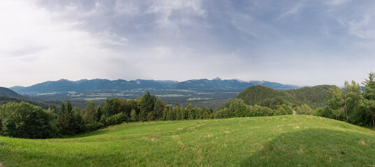 Obraz premium Wide panorama of the karawanken alps from the magical Jamnik church above Kropa in Slovenia. Blue sky and green meadows and forests in front of picturesque mountains