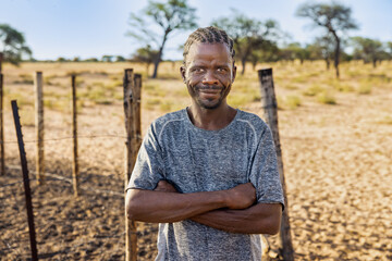 village african man with braids, standing in the yard in front of fence, sand and acacia background