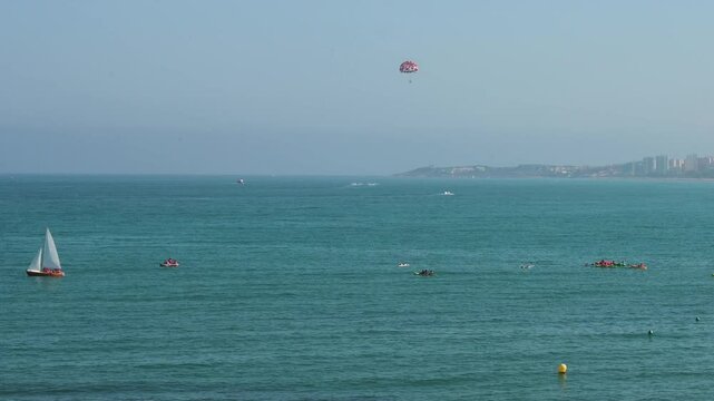 Mediterranean landscape at El Campello, Costa Blanca, Alicante, featuring a sailboat and people partaking in diverse water sports including parasailing and boating.