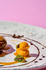 A duck confit served on a white plate with a vibrant orange sauce. The dish is garnished with fresh herbs and a drizzle of dark sauce.