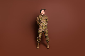 Full body photo of mature woman army soldier look empty space hands behind back wear military uniform isolated on brown color background