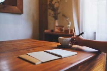 Closeup image of a hand holding a cup of coffee while writing on notebook on the table