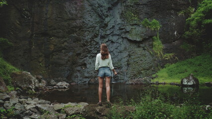 Tourist woman enjoy jungle waterfall landscape standing on rock stone. Girl relax outdoor lifestyle travel on summer holiday vacation. Picturesque scene panoramic shot. Dramatic dark toning, back view