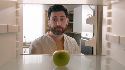 POV point of view from inside refrigerator Caucasian adult man at kitchen open empty fridge with one green apple fruit hungry guy male upset need food delivery meal crisis diet shrugs sad frustrated