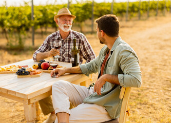 Enjoying a warm afternoon at a vineyard, two friends share stories and local wine while surrounded...