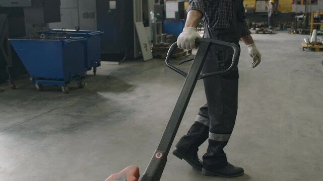 Male factory worker pushing handle of pallet jack then moving it along workshop, high angle shot