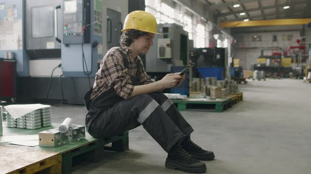 Mature Caucasian woman sitting on pallet in factory workshop and surfing Internet on smartphone during break time