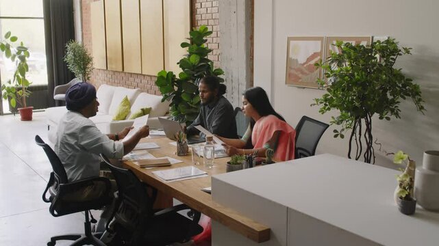Indian boss giving documents to young man and woman for research during office meeting at wooden desk