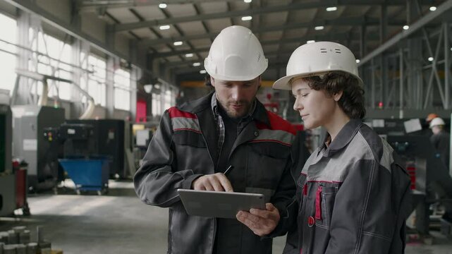 Medium shot of two engineers wearing uniform working in modern factory discussing data on digital tablet