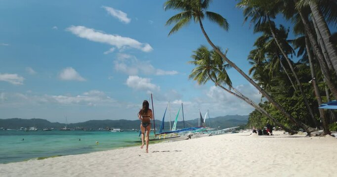 Young woman joyfully runs towards the sparkling ocean on a stunning beach with palm trees and boats, embodying the carefree spirit of a tropical island holiday in the philippines