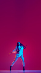Vertical image of young woman in oversized t-shirt and heeled shoes dancing against magenta studio background. Concept of hobby, entertainment, performance, creativity, lifestyle