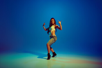 Obraz premium Passionate woman, elegant dancer in mix of sport and romantic clothes dancing on heels against blue studio background in neon light. Concept of hobby, entertainment, performance, creativity, lifestyle