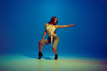 Sensual young beautiful woman dancing high heel dance, performing against blue studio background in neon light. Concept of hobby, entertainment, performance, creativity, lifestyle