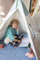 cute boy sits in a wigwam and reads a book. Reading books, children's fantasies, interesting childhood, inquisitive child, knowledge is power. Cozy interior of a children's room © Anna