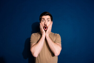 Photo of speechless man with beard dressed beige t-shirt astonished staring hold hands on cheeks isolated on dark blue color background