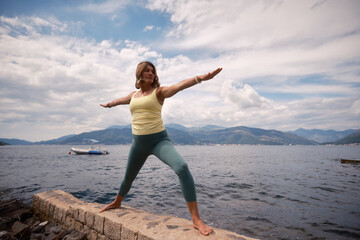 Obraz premium A woman practicing yoga in warrior pose by the serene lake under a cloudy sky