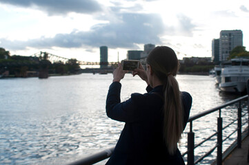 Girl taking photo with smartphone on the background of the river and the bridge