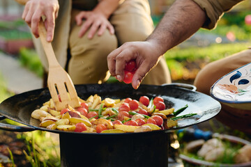 Hand of unrecognizable male farmer putting fresh cherry tomatoes on frying pan with chopped vegetables while woman mixing them