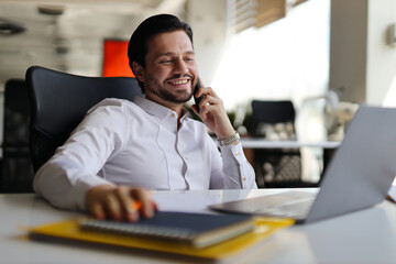 A man is talking on his cell phone while sitting at a desk. He is smiling and he is enjoying the...