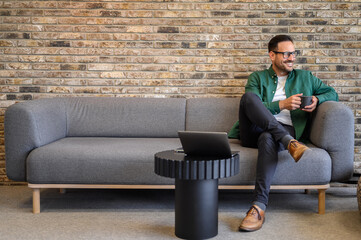 Positive male entrepreneur with coffee cup looking away thoughtfully while relaxing on sofa in...