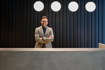 Portrait of happy handsome receptionist with arms crossed looking at camera and working at lobby...