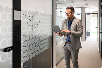 Smiling young businessman in eyeglasses working on his computer while walking in office corridor