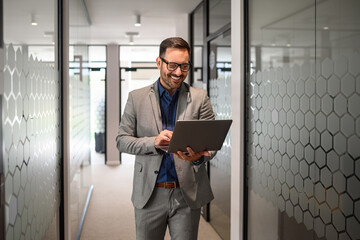 Portrait of developer smiling and doing online research over laptop while walking in office corridor