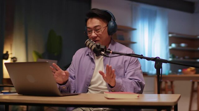 View on happy asian content creator man talking on podcast on laptop with headphones and microphone interview conversation at night broadcast studio. Home. 360 degrees. Camera moving around