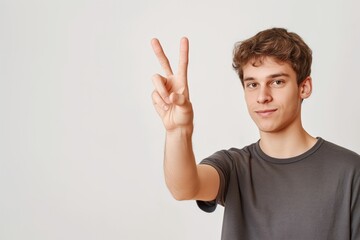 Fototapeta premium Young caucasian man with curly hair makes peace sign with right hand. Fingers forming circle. Gray t-shirt. Man face turned towards camera. Gaze directed straight at viewer. Plain white background