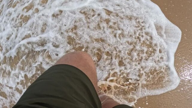 A person is walking on the beach with their feet in the sand. The sand is wet and the person's feet are visible