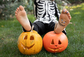 child sitting on the grass with his bare feet on carved orange pumpkins with a Halloween grin. Preparing for Halloween. cheerful childhood, pampering. Don't be afraid, have fun