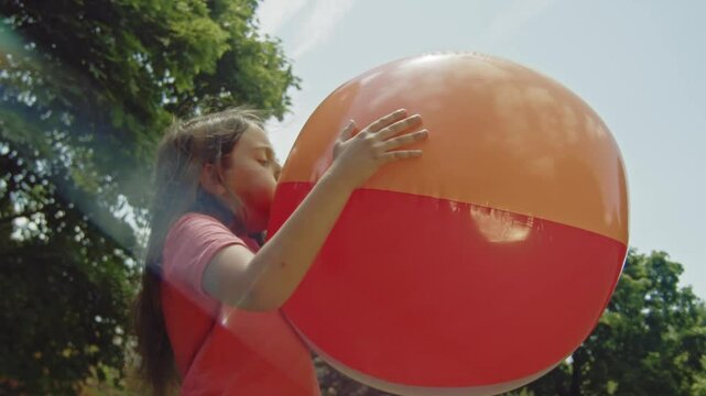 A young girl is seen inflating a large beach ball in the park, soaking up the sun on a warm summer day. Her focus and enthusiasm reflect the fun of summer activities.