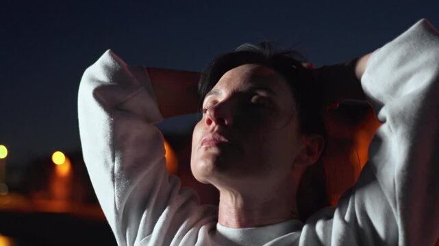 Thoughtful sporty woman looking away in city at night. Young fit female with hands behind head is looking at camera in the dark outdoors. 