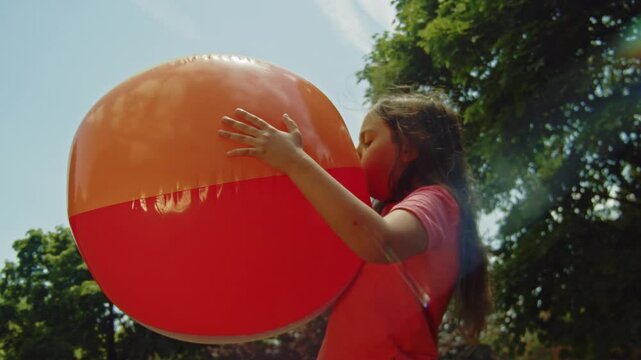 A cheerful girl works on inflating a big beach ball in the park, taking advantage of the beautiful summer weather. The scene captures the simple joys of outdoor playtime.