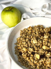 Granola with nuts and raisins in a white plate with apple close-up on a white textile background. Top view. Healthy proper breakfast.
