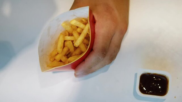The man's hands take French fries from a red paper package. He dips a piece into the sauce and eats it. Top view of fast food and a white table in a cafe